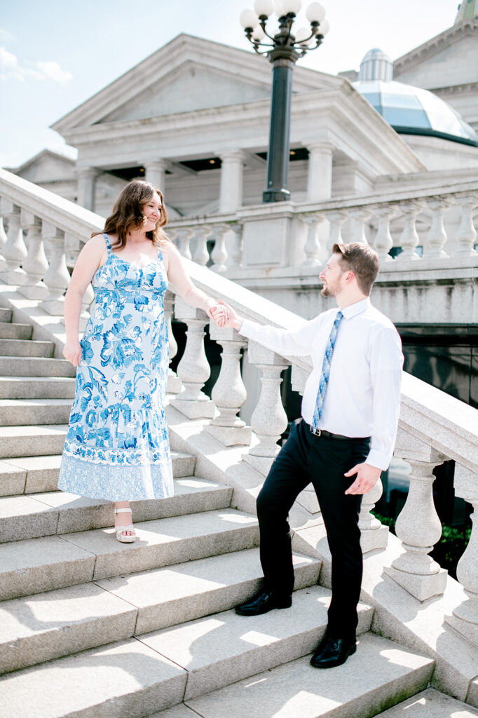 Man and woman hold hands on staircase with pillars. The woman is in a blue and white patterned dress.