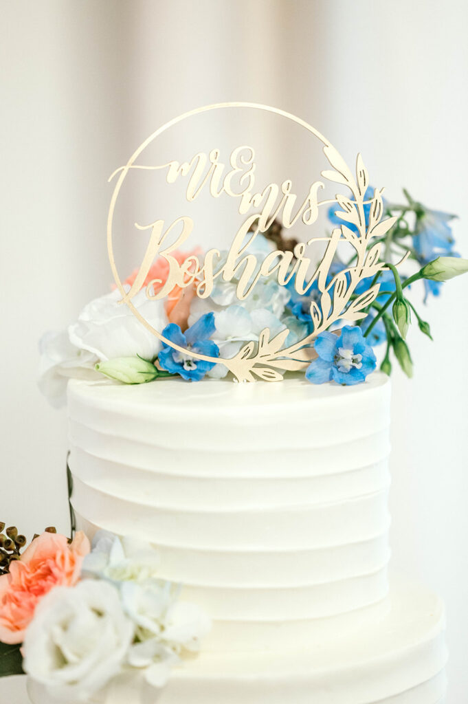 Wedding day essential cake sits on a table. It has white frosting. Around the top and bottom there are blue, white, and orange candy flowers.